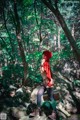 A woman in a red hoodie standing on a rock in the woods.