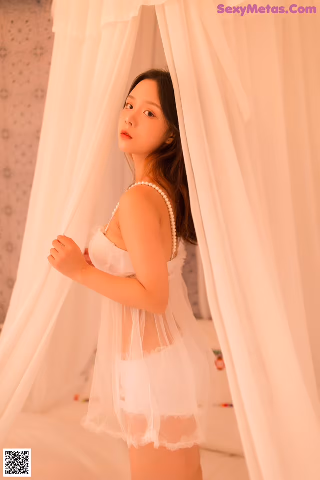 A woman in a white lingerie standing in front of a curtain.